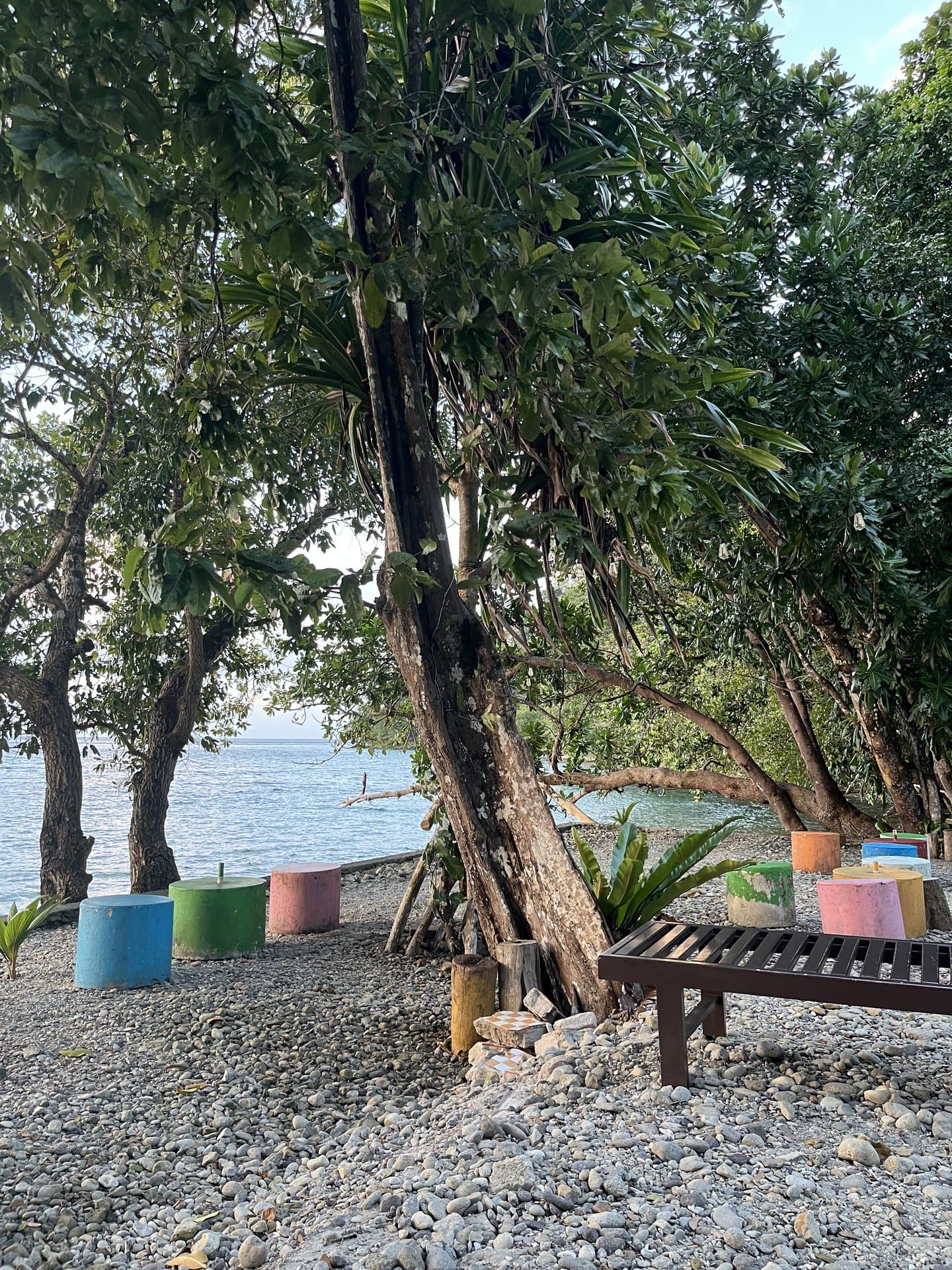Tepi Pantai Memit Shafira Resort — pantai berkerikil rindang dengan bangku santai dan laut jernih